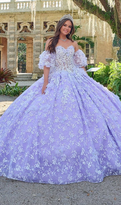 A young woman in the Quinceanera Dress 54206 By Amarra, a lavender ball gown with floral lace details, stands smiling outdoors in front of an ornate stone building.