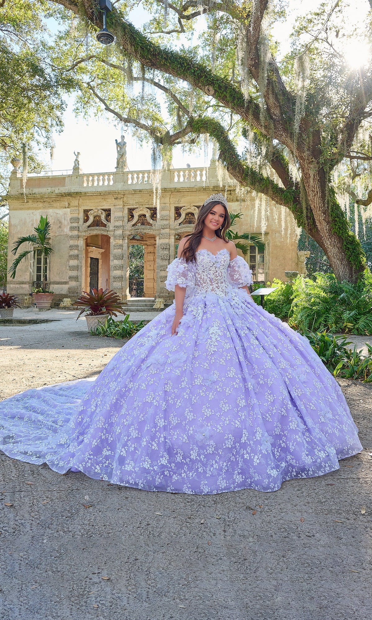A young woman stands outdoors in front of a historic building, wearing the Quinceanera Dress 54206 By Amarra—a lavender ball gown with white floral details and an off-the-shoulder design.