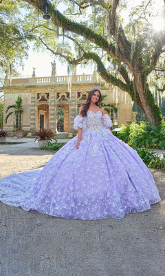 A young woman stands outdoors in front of a historic building, wearing the Quinceanera Dress 54206 By Amarra—a lavender ball gown with white floral details and an off-the-shoulder design.