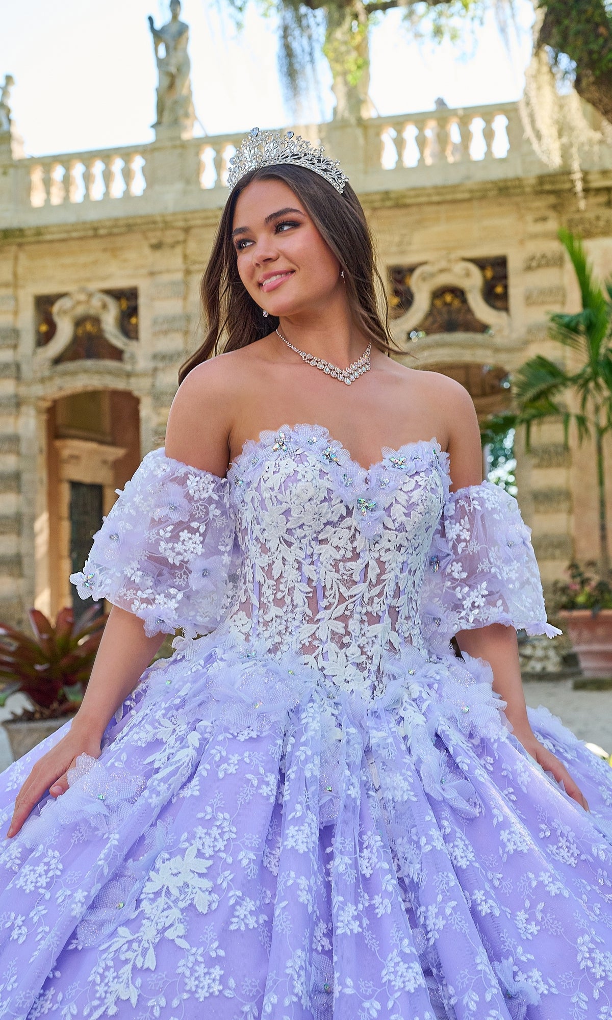 A young woman in the Quinceanera Dress 54206 By Amarra, a lavender ball gown with glow-in-the-dark floral lace and a tiara, stands outdoors before a stone building.