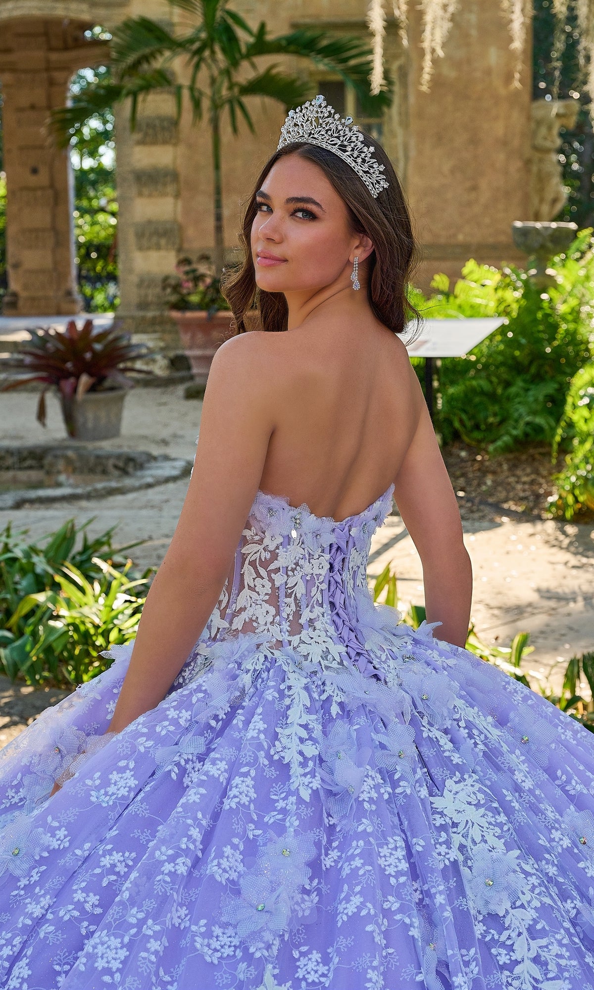 A young woman poses outdoors, looking over her shoulder in the Quinceanera Dress 54206 By Amarra, a ball gown adorned with white floral details and a tiara.