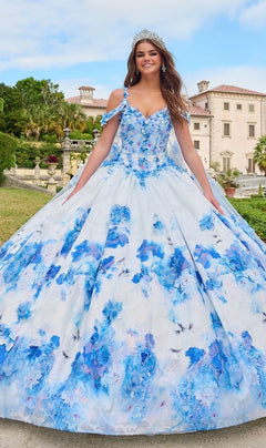 A young woman wearing the Quinceanera Dress 54208 By Amarra, a blue and white floral ball gown with a tiara, stands outdoors in front of a grand building.