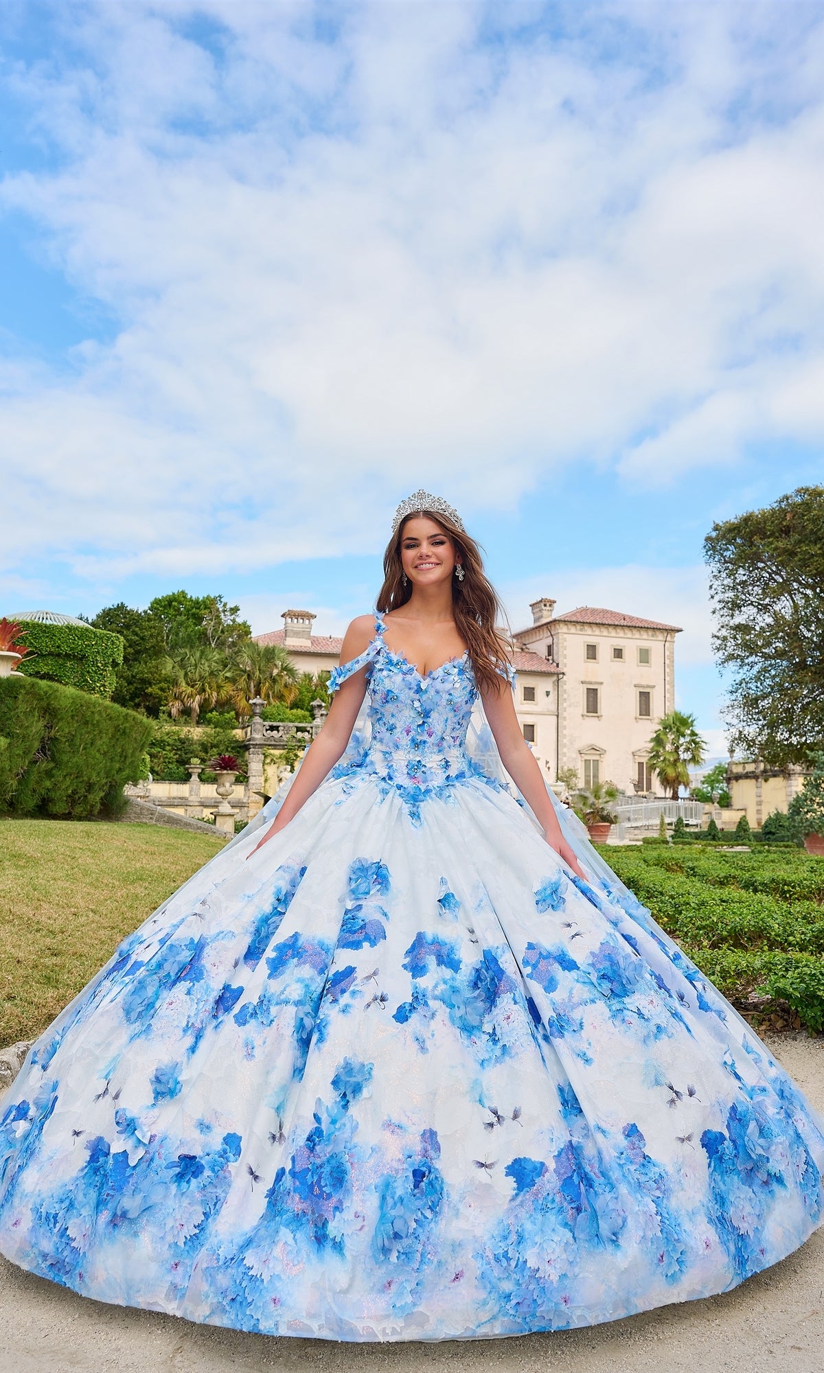 Wearing the Quinceanera Dress 54208 by Amarra, a young woman in a blue and white floral ball gown stands smiling outside a grand mansion on a sunny day, topped with a sparkling tiara.