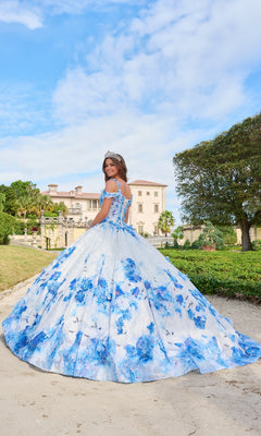 A young woman wearing the Quinceanera Dress 54208 By Amarra, featuring floral prints and glitter tulle, stands on a path outdoors with a mansion and lush greenery in the background under a partly cloudy sky.