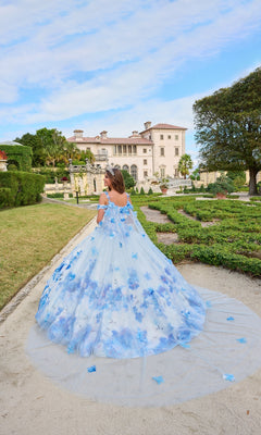 A woman stands outdoors on a garden path, facing a mansion, wearing the Quinceanera Dress 54208 By Amarra in blue and white floral print.