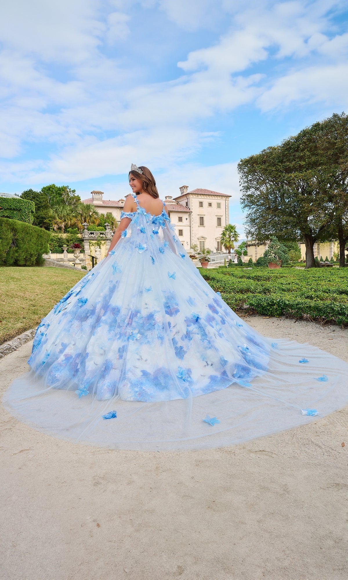 A woman, seen from behind, wears the Quinceanera Dress 54208 by Amarra—a flowing blue and white ball gown—while standing outdoors before a grand historic building and manicured gardens.