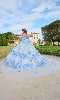A woman, seen from behind, wears the Quinceanera Dress 54208 by Amarra—a flowing blue and white ball gown—while standing outdoors before a grand historic building and manicured gardens.