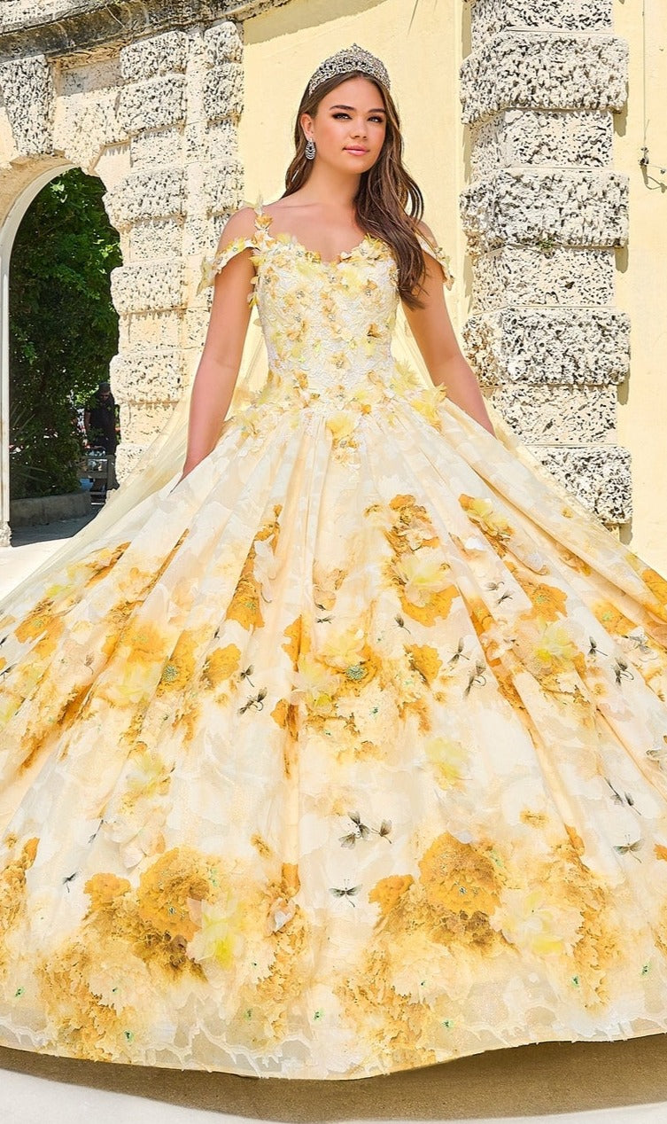 A young woman in the Quinceanera Dress 54208 by Amarra, a yellow floral-print ball gown with a tiara, stands outdoors before a stone archway.