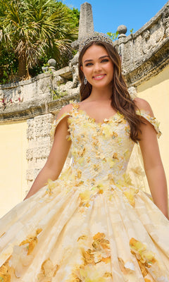 A young woman in the Quinceanera Dress 54208 by Amarra—a floral print ball gown with glitter tulle and a jeweled tiara—smiles at the camera while standing outdoors in front of a stone wall and palm trees.