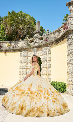 A young woman in the Quinceanera Dress 54208 By Amarra poses outdoors against an ornate stone wall and greenery beneath a clear blue sky.