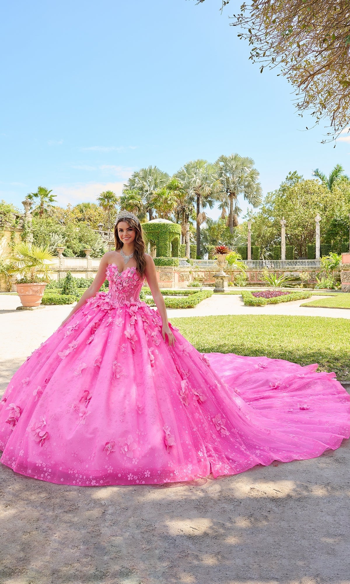 Outdoors in a garden, a young woman wears the Quinceanera Dress 54218 by Amarra—a bright pink ball gown with puff sleeves, floral details, tiara, and a long glitter tulle train cascading behind her.