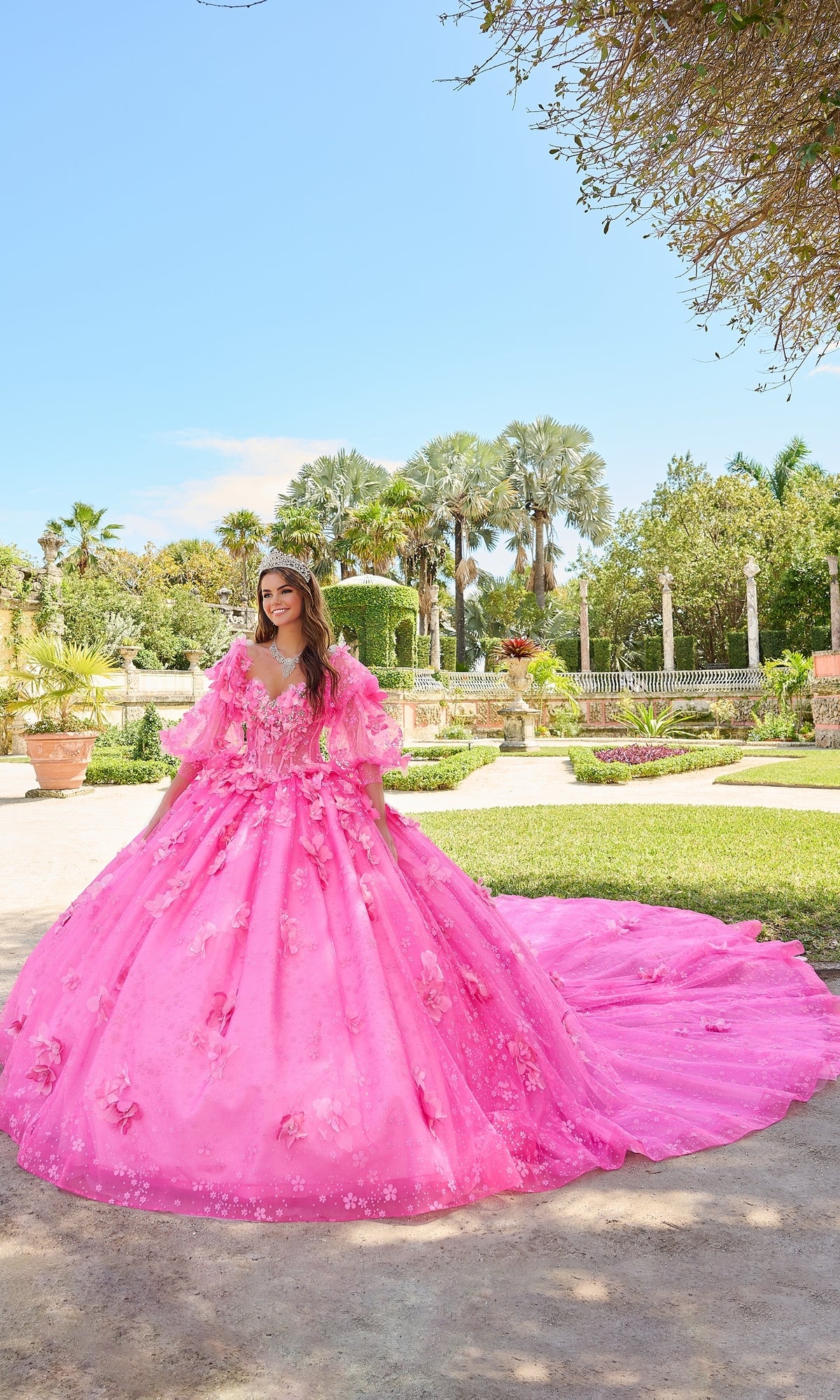 A young woman stands in a garden wearing Quinceanera Dress 54218 By Amarra, featuring bright pink color, puff sleeves, floral details, and a glitter tulle long train.