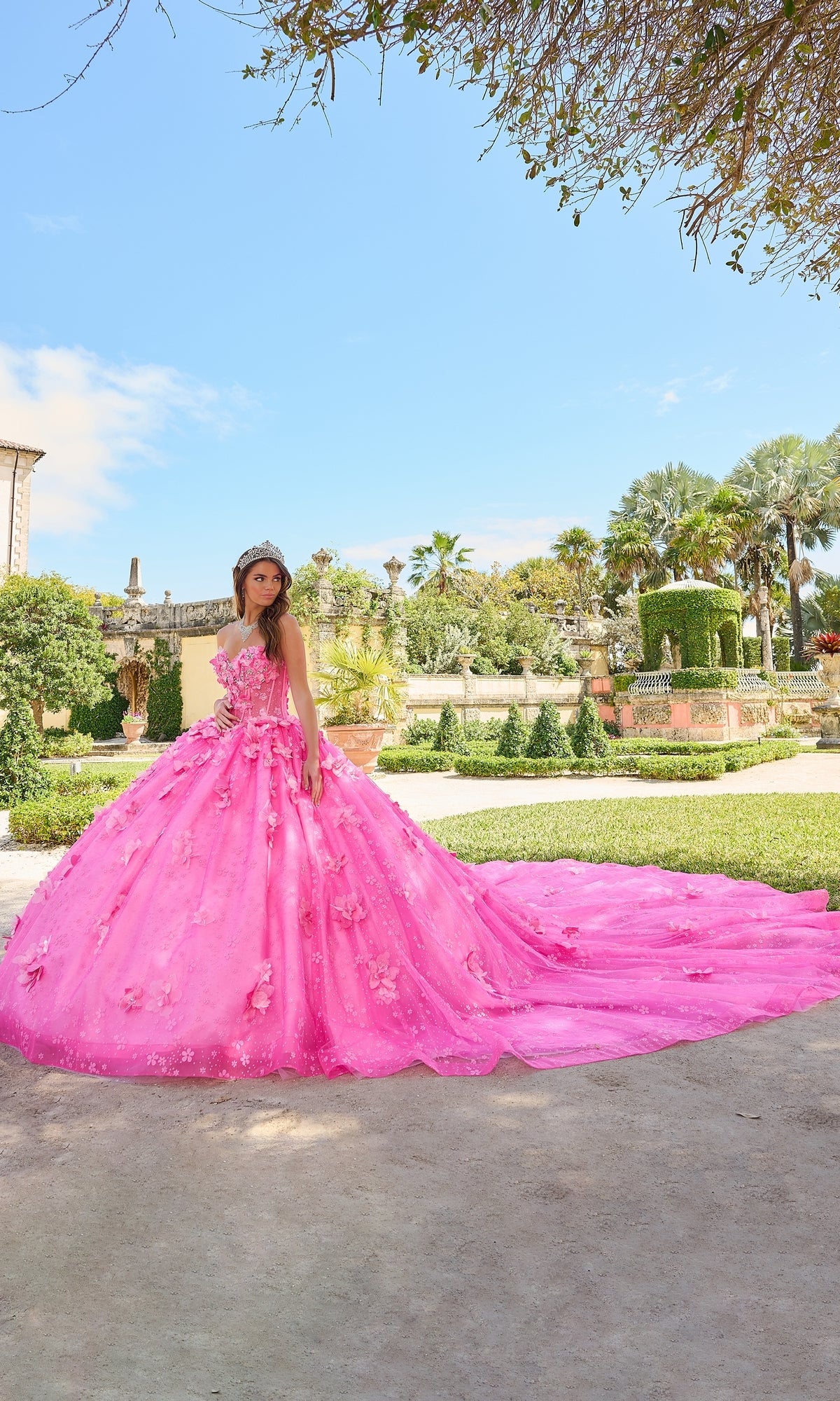 A young woman stands outside in a garden wearing the Quinceanera Dress 54218 By Amarra, a bright pink ball gown featuring puff sleeves, floral details, glitter tulle, and a sparkling tiara.