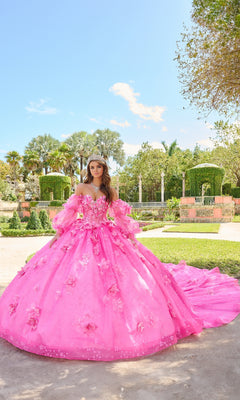 A young woman poses in a garden wearing the Quinceanera Dress 54218 By Amarra, featuring glitter tulle, puff sleeves, floral details, and a tiara for a formal portrait.