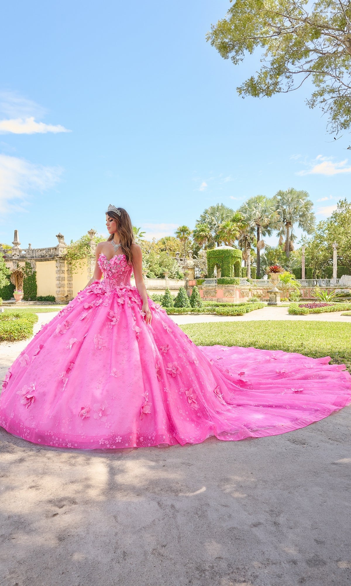 A person wears the Quinceanera Dress 54218 By Amarra, a bright pink ball gown with puff sleeves and floral details, standing outdoors in a landscaped garden under a sunny sky.