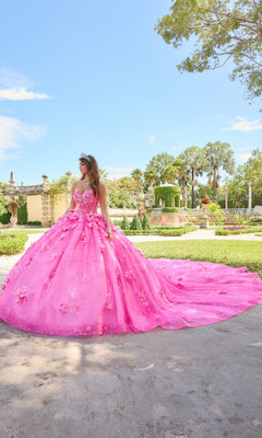 A person wears the Quinceanera Dress 54218 By Amarra, a bright pink ball gown with puff sleeves and floral details, standing outdoors in a landscaped garden under a sunny sky.