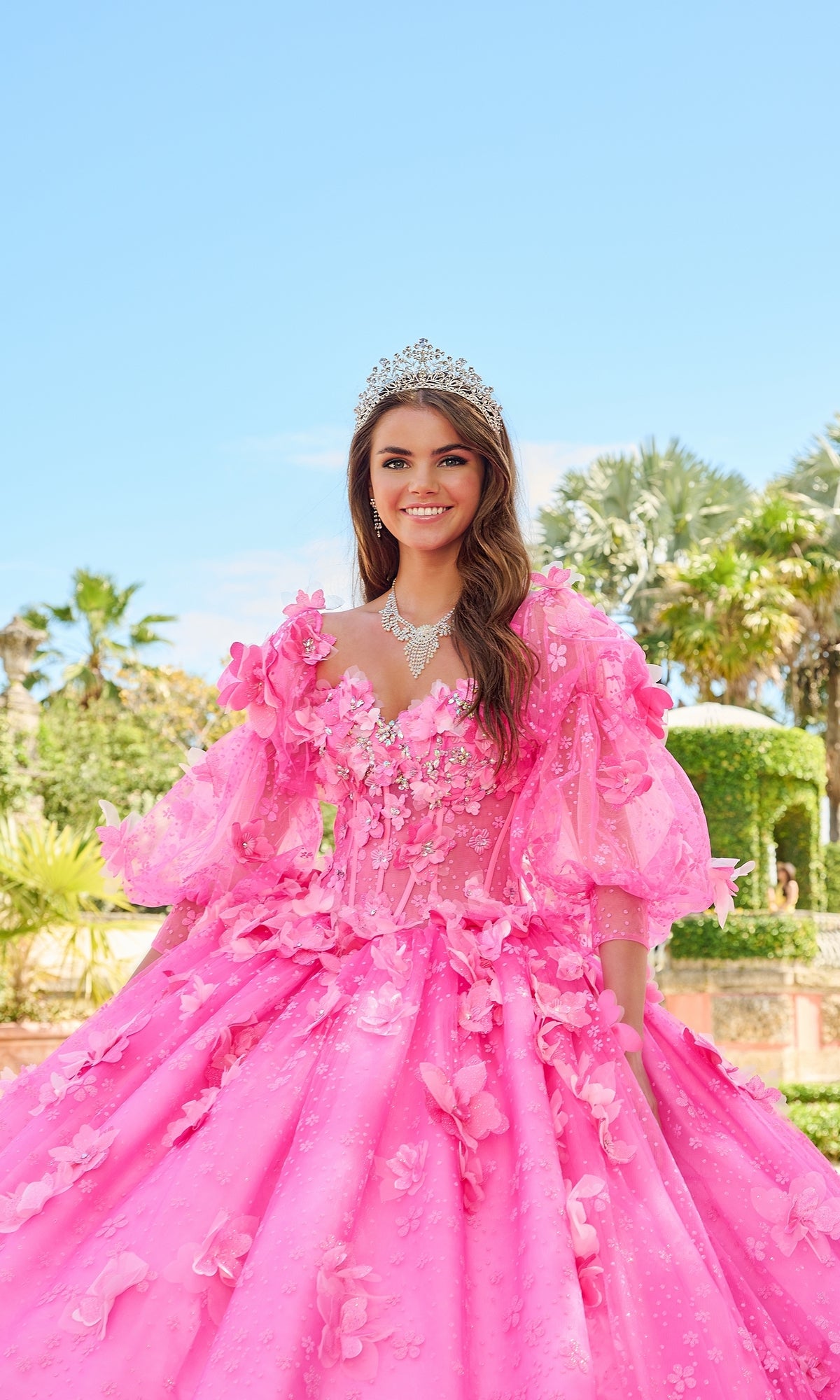 A young woman smiles outdoors among trees and hedges, wearing the Quinceanera Dress 54218 By Amarra—a bright pink ball gown with puff sleeves and a tiara.