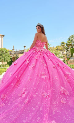 A young woman wears the Quinceanera Dress 54218 by Amarra, a vibrant pink glitter tulle ball gown with puff sleeves and a tiara, as she stands outdoors in a garden facing slightly away from the camera.