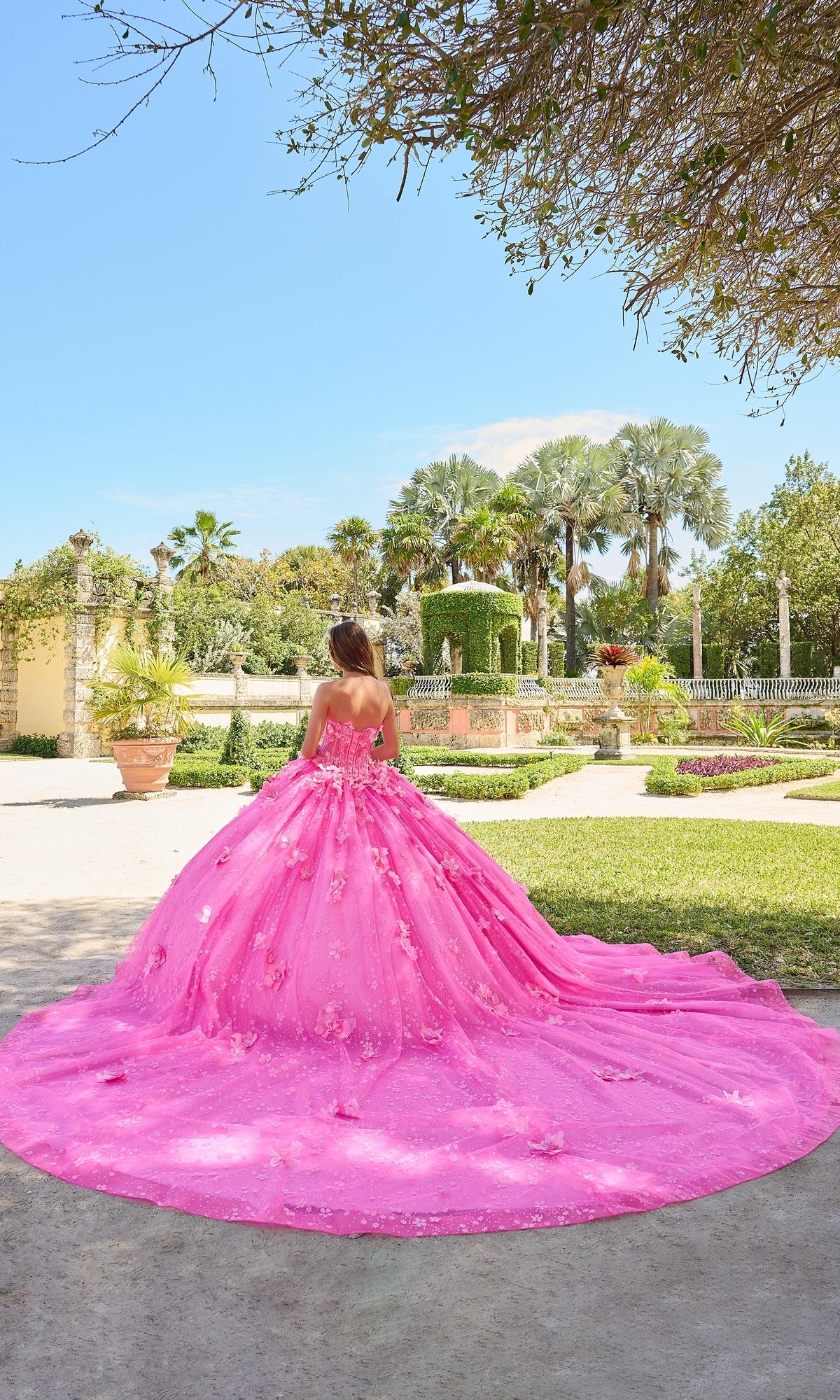 A person stands outdoors in a landscaped garden under a clear blue sky, wearing the Quinceanera Dress 54218 By Amarra, a bright pink ball gown with puff sleeves.