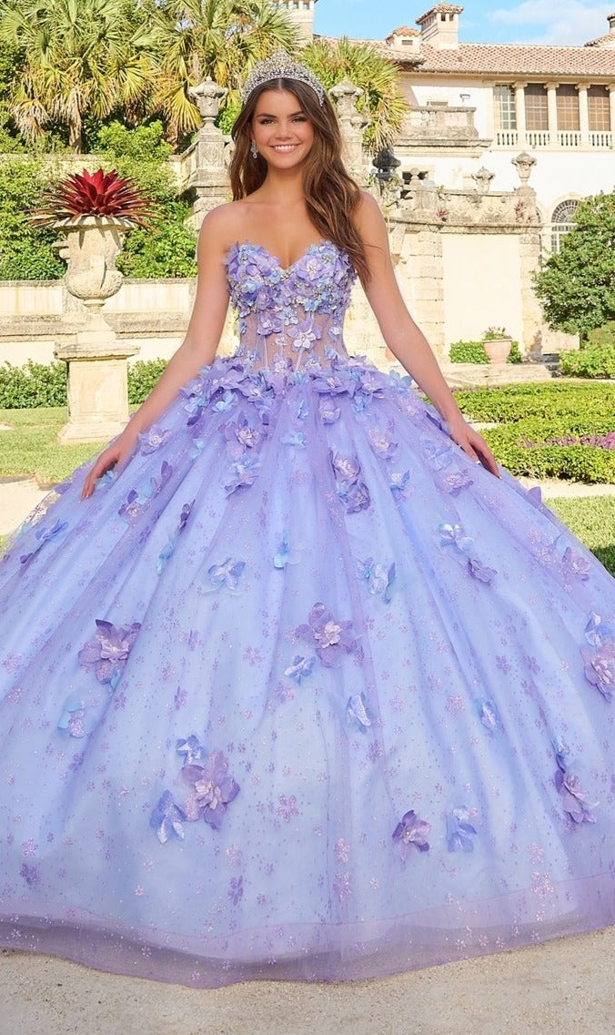 A woman stands outdoors in the Quinceanera Dress 54218 by Amarra, a strapless lavender ball gown featuring floral appliqués and glitter tulle, its voluminous skirt elegantly fanned out around her.