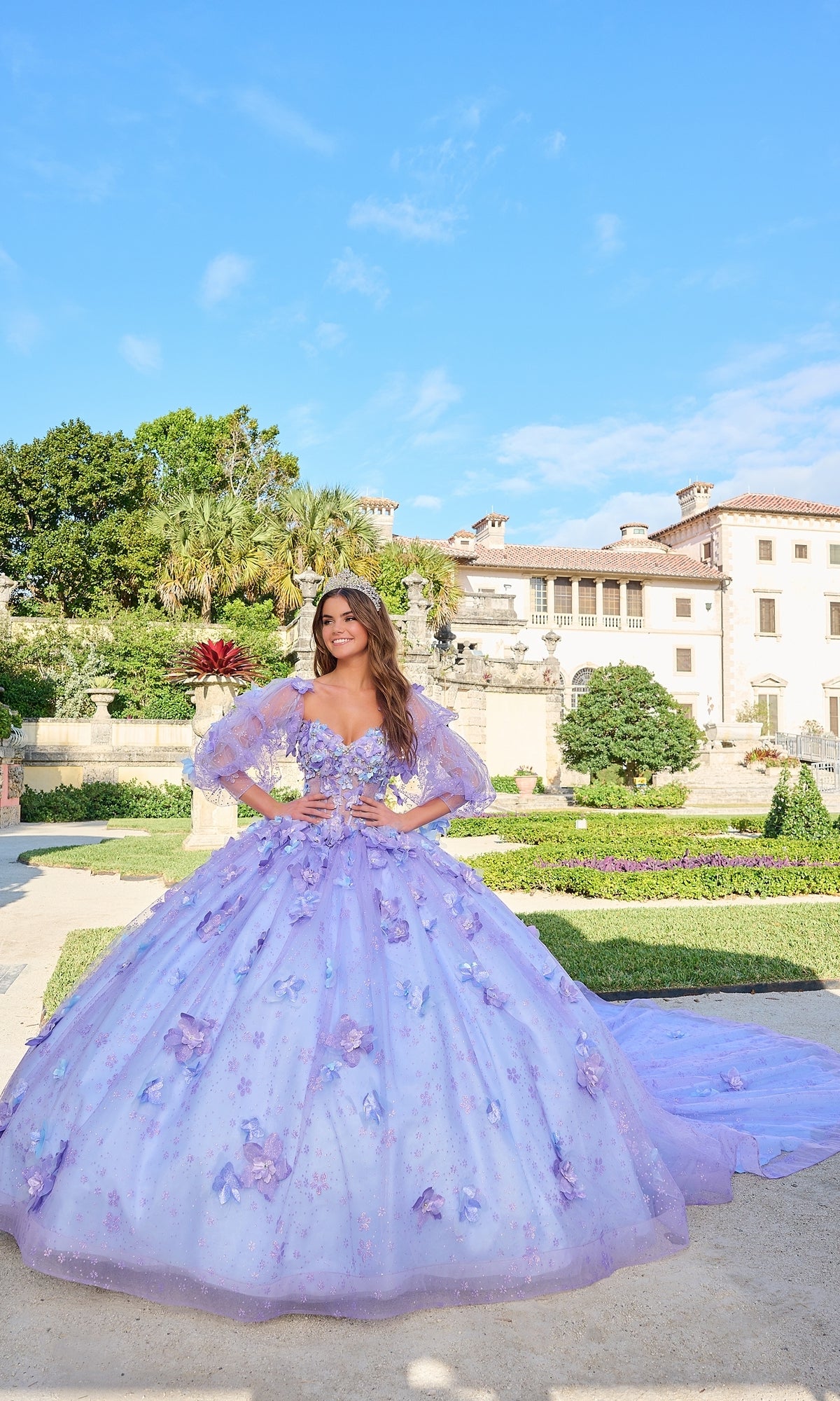 A woman wears the Quinceanera Dress 54218 by Amarra, featuring floral details and puff sleeves, as she stands in a formal garden before a grand historic building under a clear blue sky.