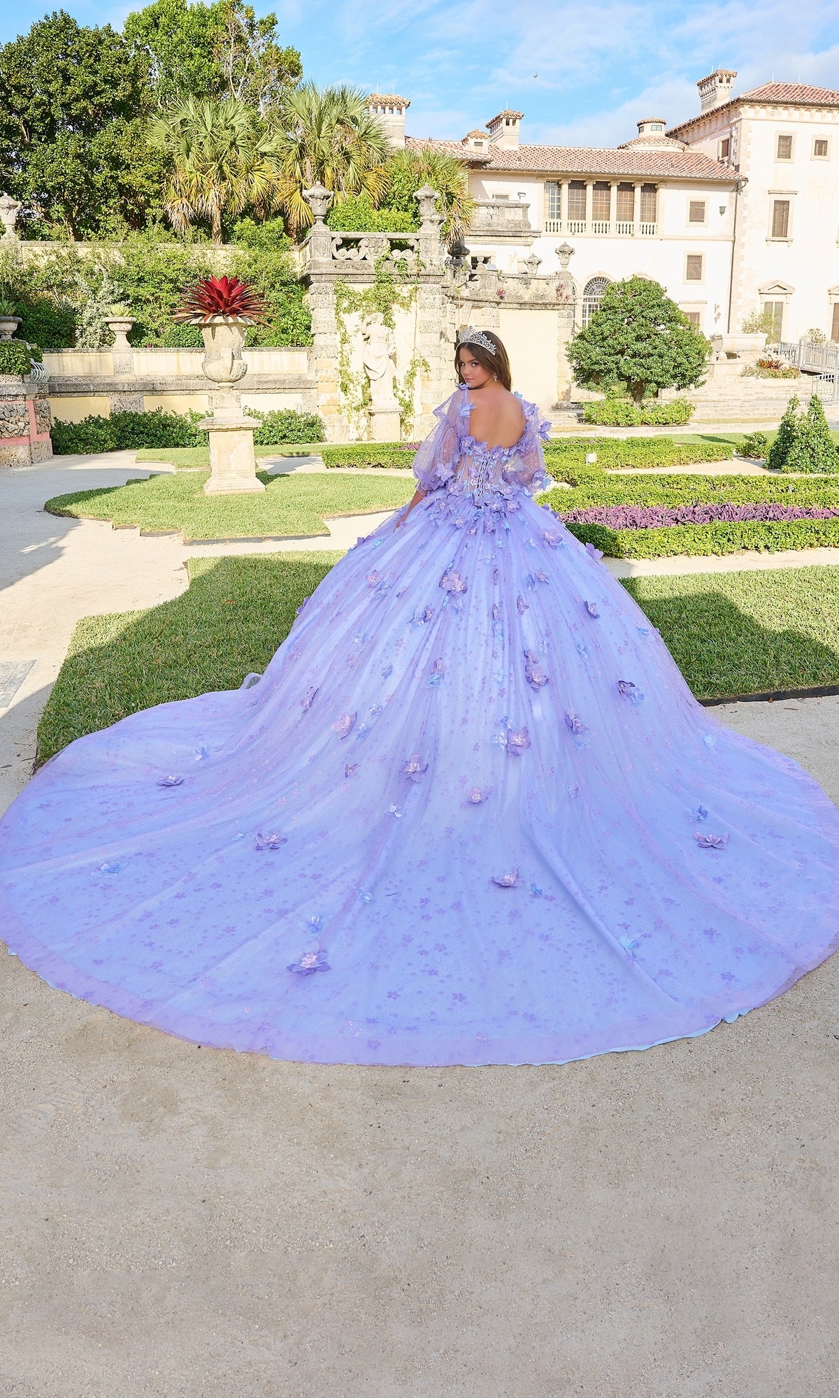 A woman wearing the Quinceanera Dress 54218 By Amarra, a voluminous lavender ball gown with floral details and elegant puff sleeves, stands in a formal garden with a mansion in the background.