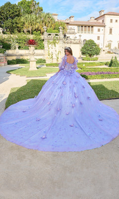 A woman wearing the Quinceanera Dress 54218 By Amarra, a voluminous lavender ball gown with floral details and elegant puff sleeves, stands in a formal garden with a mansion in the background.