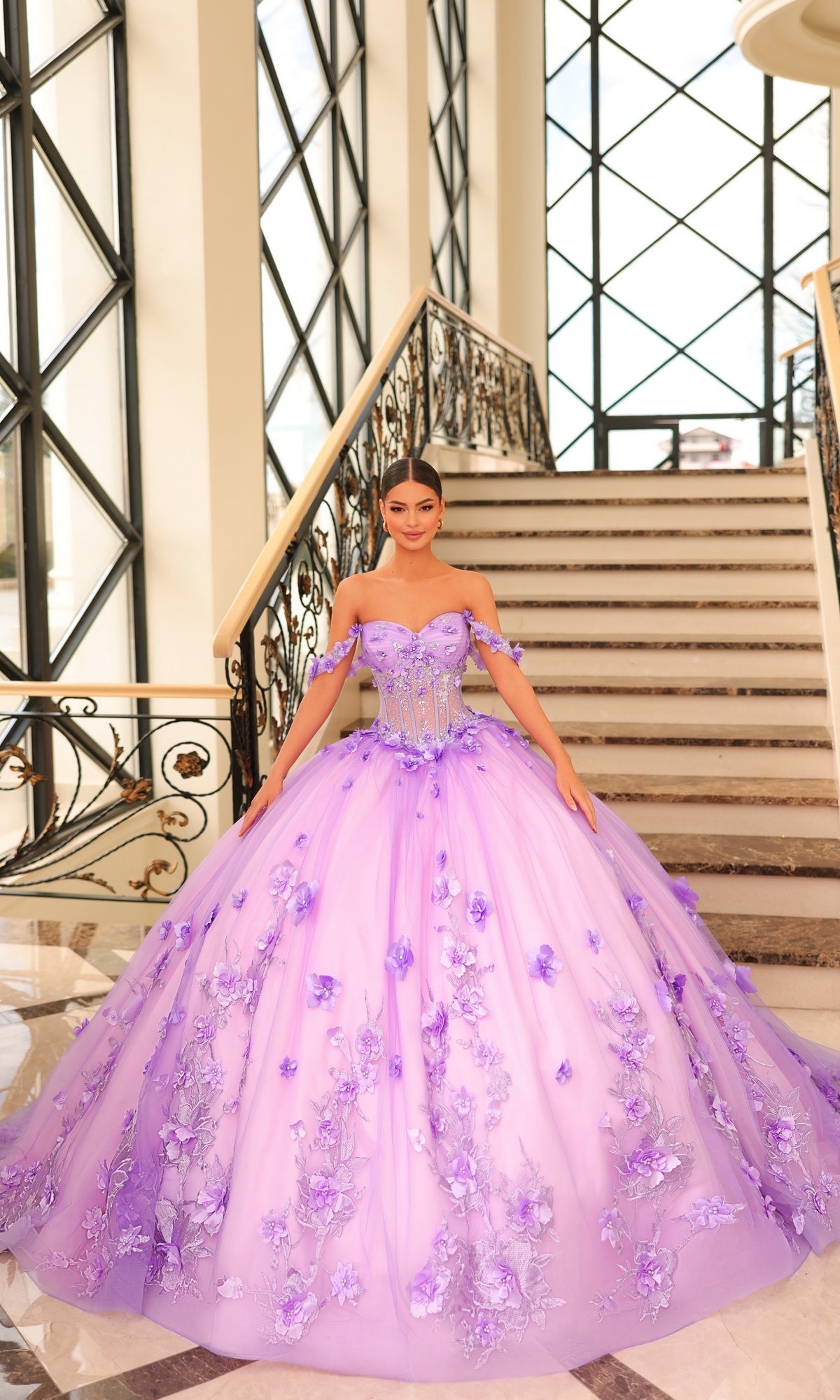 A woman stands on marble stairs wearing the Quinceanera Dress 54224 by Amarra, a voluminous lavender tulle ball gown with floral embellishments and off-the-shoulder sleeves.
