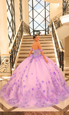 A young woman in the Quinceanera Dress 54224 By Amarra, a lavender tulle ball gown, stands at the base of a grand staircase with wrought iron railings and large windows in the background.