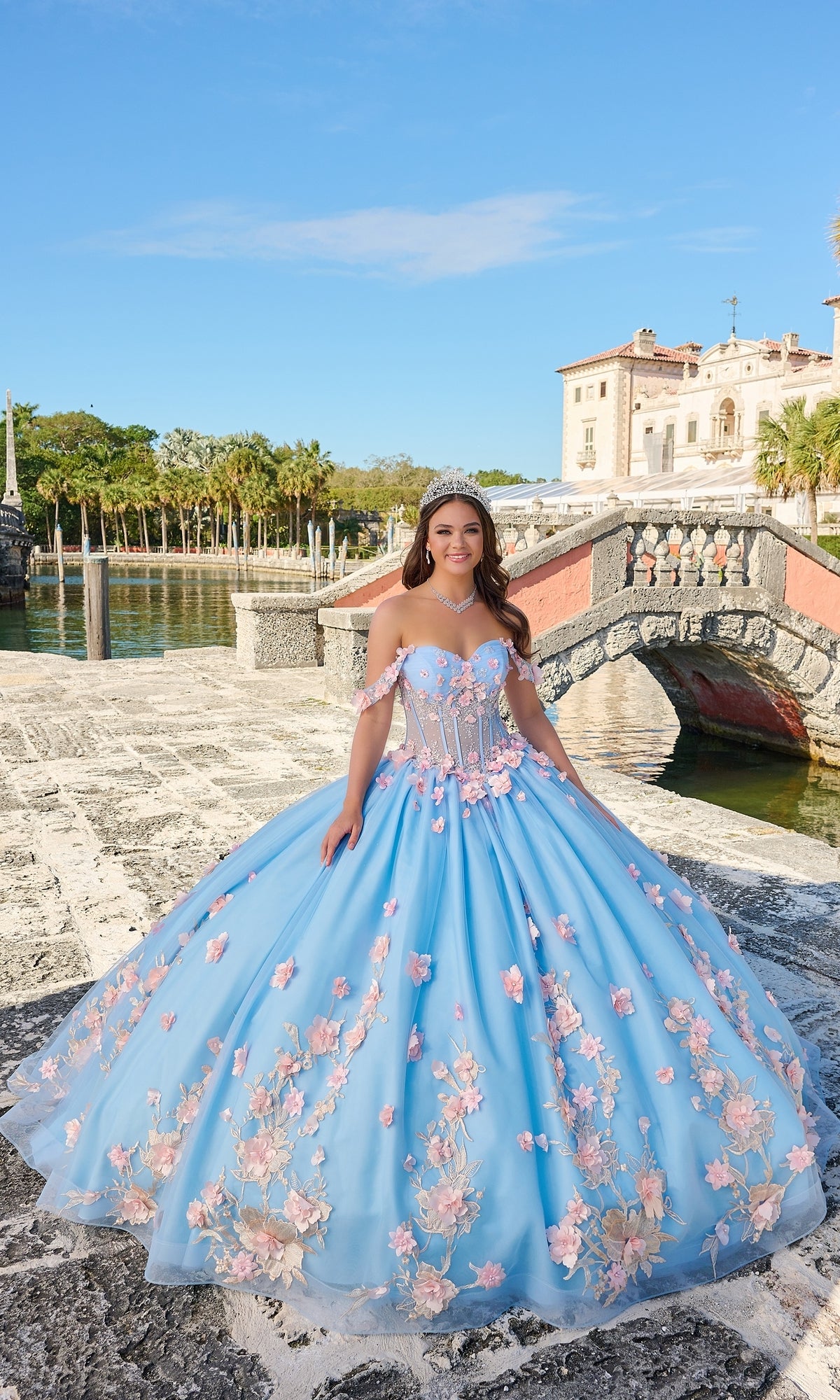 A young woman in the Quinceanera Dress 54224 By Amarra, a light blue tulle ball gown with pink floral appliqué, stands outdoors on a stone walkway near a bridge and water, with a historic building behind her.
