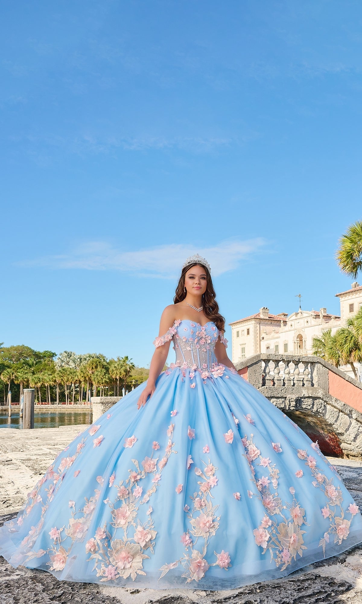 A young woman wears the Quinceanera Dress 54224 by Amarra, a blue off-the-shoulder tulle gown with pink floral appliques, standing outdoors near a mansion, palm trees, and a stone bridge on a sunny day.