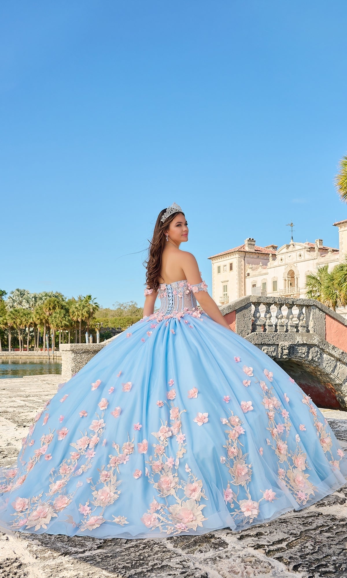A young woman stands outdoors near a historic stone building, wearing the Quinceanera Dress 54224 By Amarra—a blue tulle ball gown with pink floral appliqué and a tiara—under a clear blue sky.