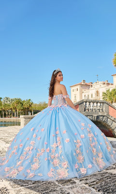 A young woman stands outdoors near a historic stone building, wearing the Quinceanera Dress 54224 By Amarra—a blue tulle ball gown with pink floral appliqué and a tiara—under a clear blue sky.