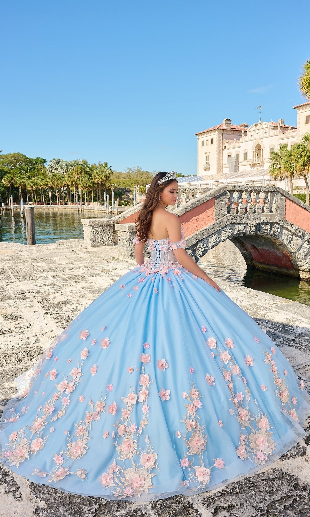 A young woman in the Quinceanera Dress 54224 By Amarra, featuring off-the-shoulder pink floral appliqué and a tiara, stands on a stone pathway by water and a historic building.