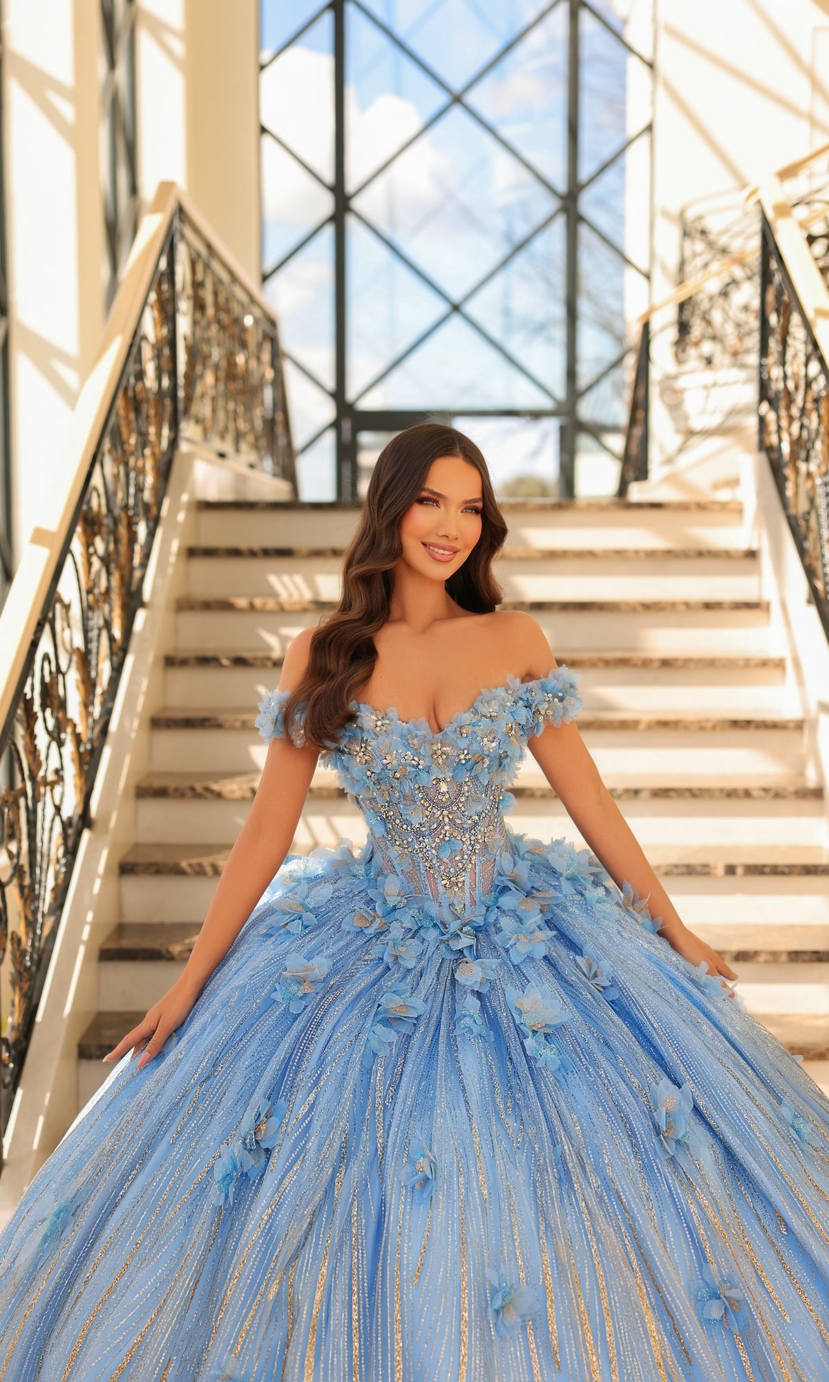 A woman wearing the Quinceanera Dress 54226 By Amarra, a blue off-the-shoulder ball gown with floral applique, smiles on a staircase with ornate railings and large windows in the background.