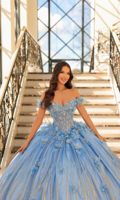 A woman wearing the Quinceanera Dress 54226 By Amarra, a blue off-the-shoulder ball gown with floral applique, smiles on a staircase with ornate railings and large windows in the background.