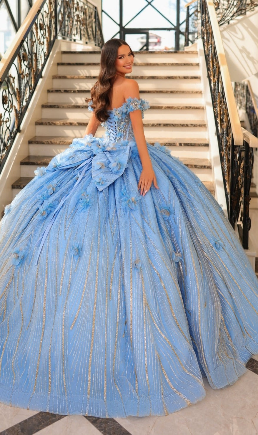 A woman in the Quinceanera Dress 54226 by Amarra, an off-the-shoulder sparkling blue ball gown, stands on a staircase and looks over her shoulder with her hand on the railing.