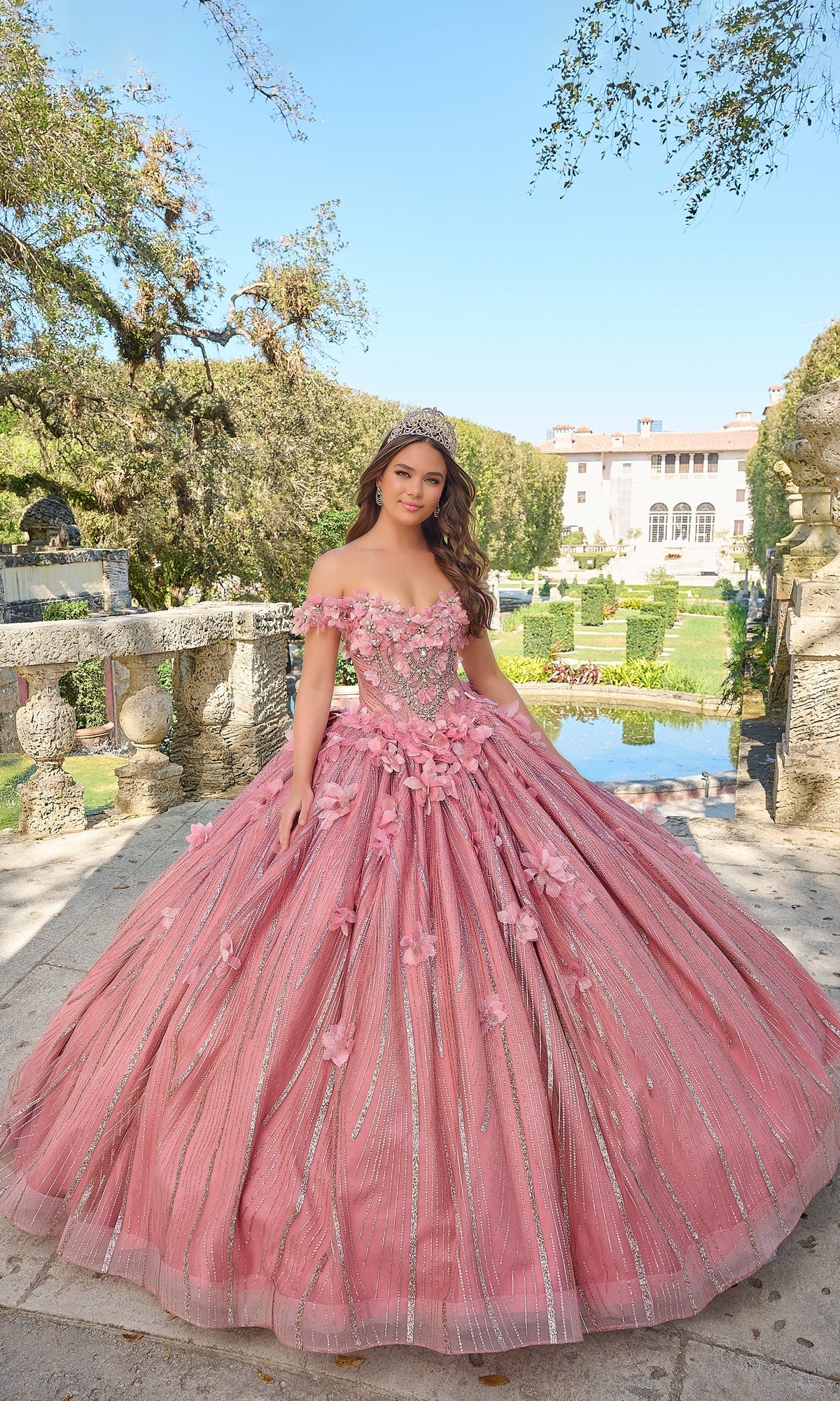 A young woman stands outdoors before a garden and grand estate wearing the Quinceanera Dress 54226 By Amarra, a pink off-the-shoulder ball gown adorned with intricate floral appliqué.