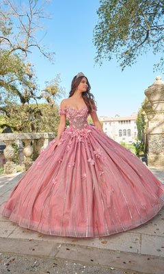A young woman wearing the Quinceanera Dress 54226 by Amarra, featuring off-the-shoulder floral appliqué and a tiara, stands outdoors on stone steps in front of a historic building surrounded by greenery.