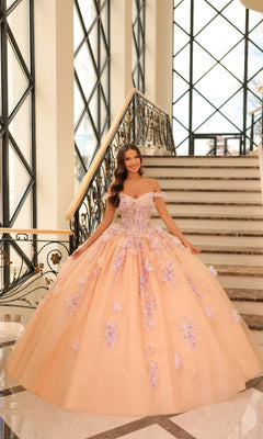 A woman in the Quinceanera Dress 54228 By Amarra, a peach ball gown with 3D floral appliqué and an elegant off-the-shoulder design, stands smiling at the base of a grand staircase bathed in sunlight from large windows.