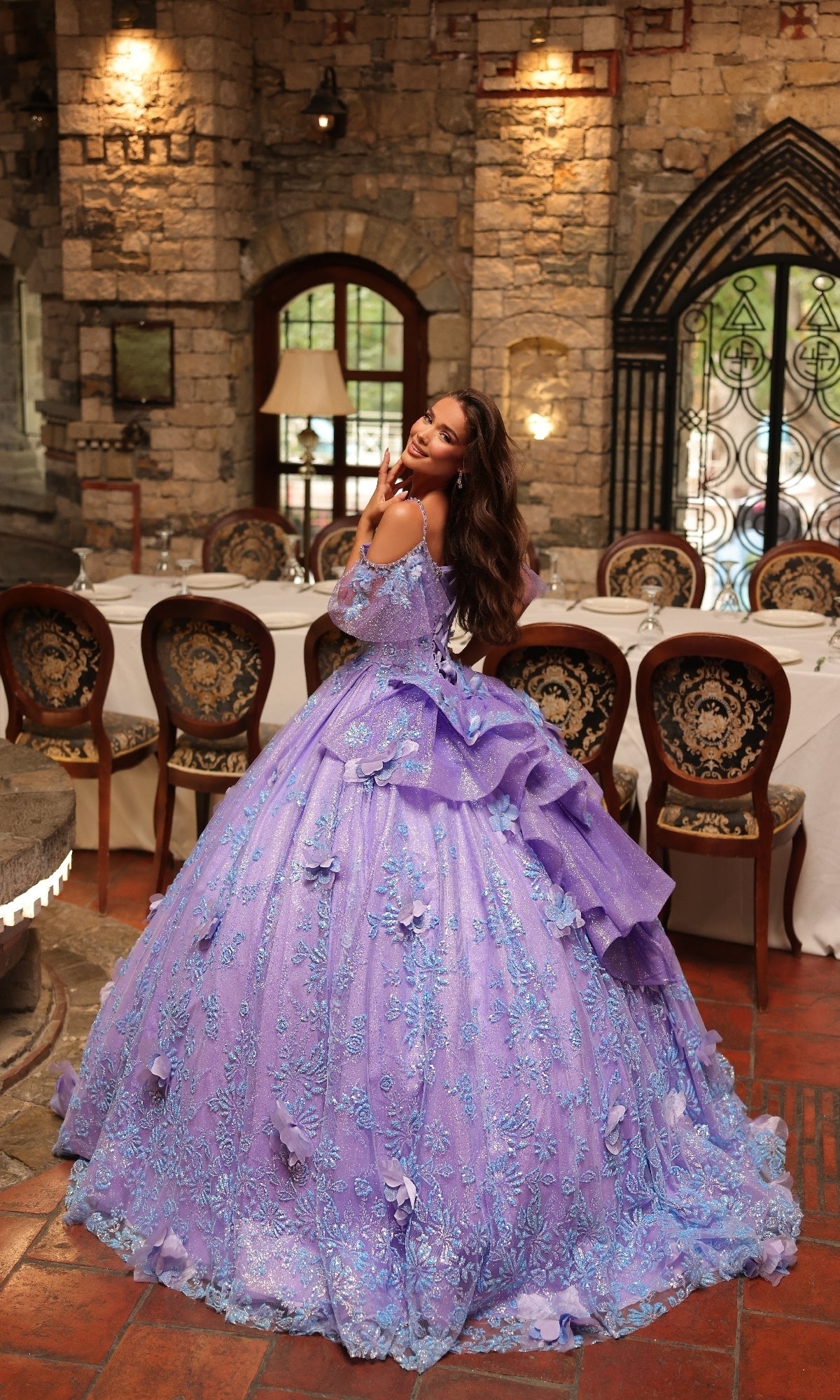 A woman wearing the Amarra 54229 Glitter Tulle Quinceañera Dress stands in an elegant stone-walled dining room with a long table and upholstered chairs.