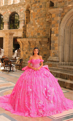 A young woman wears the Amarra 54233, a bright pink 3D Flower Quinceanera Dress with detachable straps, standing outside a stone building with arched doorways and nearby empty chairs.