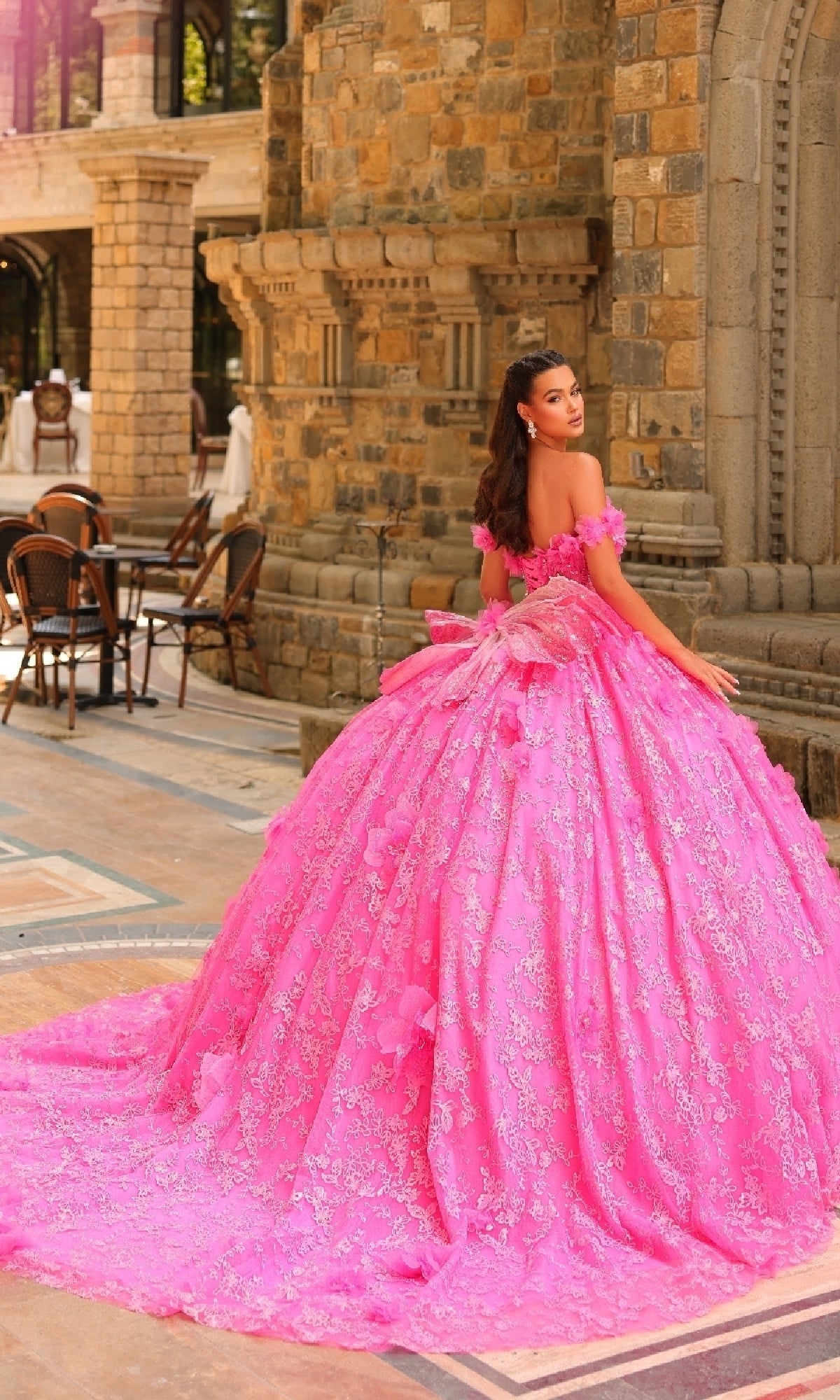 A woman in the Amarra 54233 3D Flower Quinceanera Dress with detachable straps stands outdoors by stone architecture, looking over her shoulder.