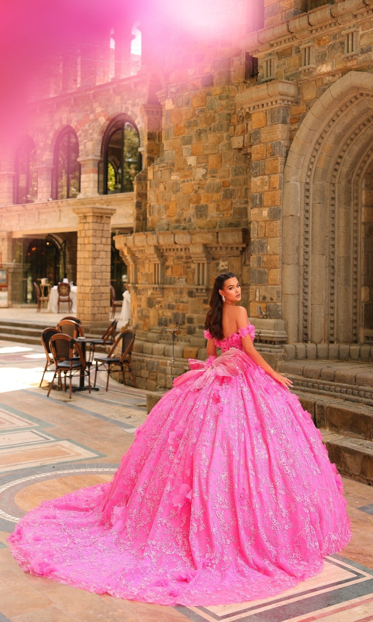 A woman in the Amarra 54233 3D Flower Quinceanera Dress stands on a patterned stone courtyard before a historic building with arched windows.