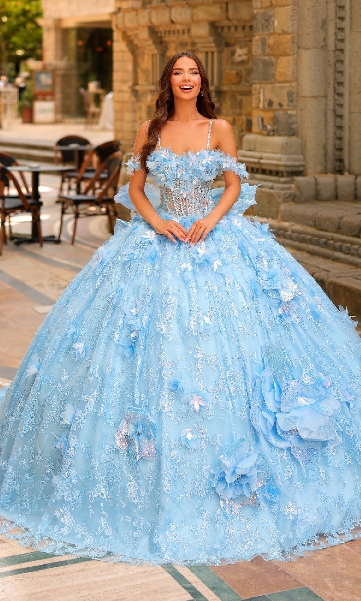 A woman stands outdoors in the Amarra 54233 3D Flower Quinceanera Dress, a voluminous light blue ball gown adorned with lace, 3D flowers, and elegant floral details.