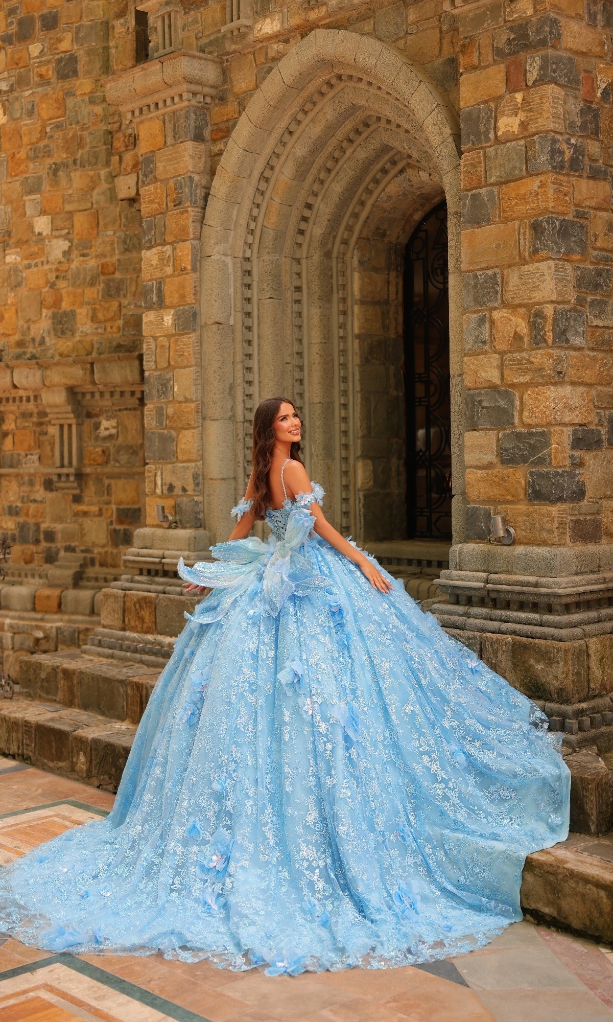 A woman in the Amarra 54233 3D Flower Quinceanera Dress poses in front of a grand stone arched doorway, glancing over her shoulder.