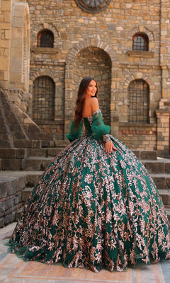 A woman stands outdoors in front of a stone building, wearing the Sequin Quinceanera Dress: Amarra 54238, an off-shoulder green ball gown with gold embroidery, a corset bodice, and a voluminous skirt.