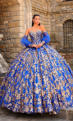 A woman stands in front of a stone building wearing the Sequin Quinceanera Dress: Amarra 54238, a blue ball gown featuring gold embellishments, a corset bodice, and sheer blue sleeves.