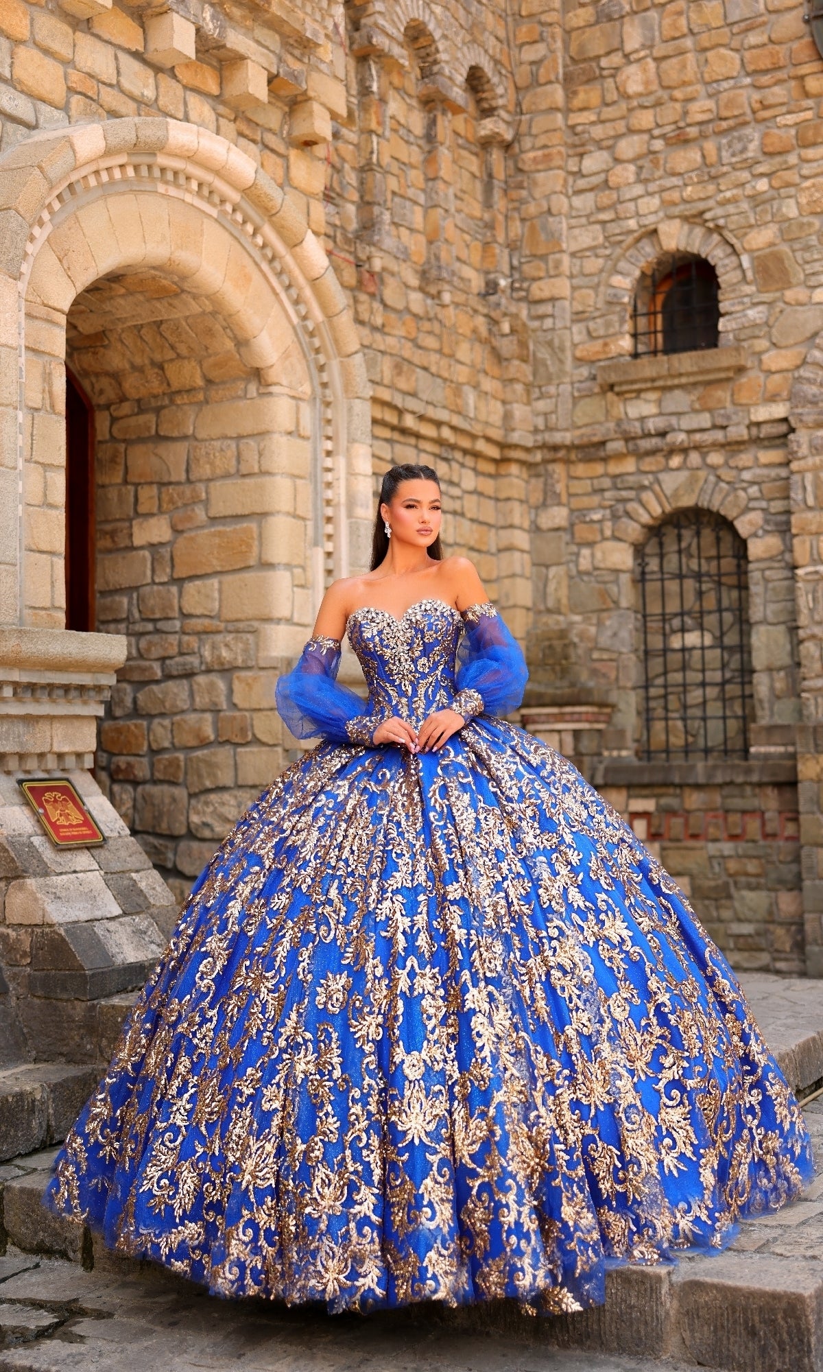 A woman stands before a stone building wearing the Sequin Quinceanera Dress: Amarra 54238, featuring an ornate blue ball gown design with gold embroidery, a corset bodice, and matching sheer sleeves.