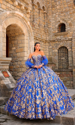 A woman stands before a stone building wearing the Sequin Quinceanera Dress: Amarra 54238, featuring an ornate blue ball gown design with gold embroidery, a corset bodice, and matching sheer sleeves.
